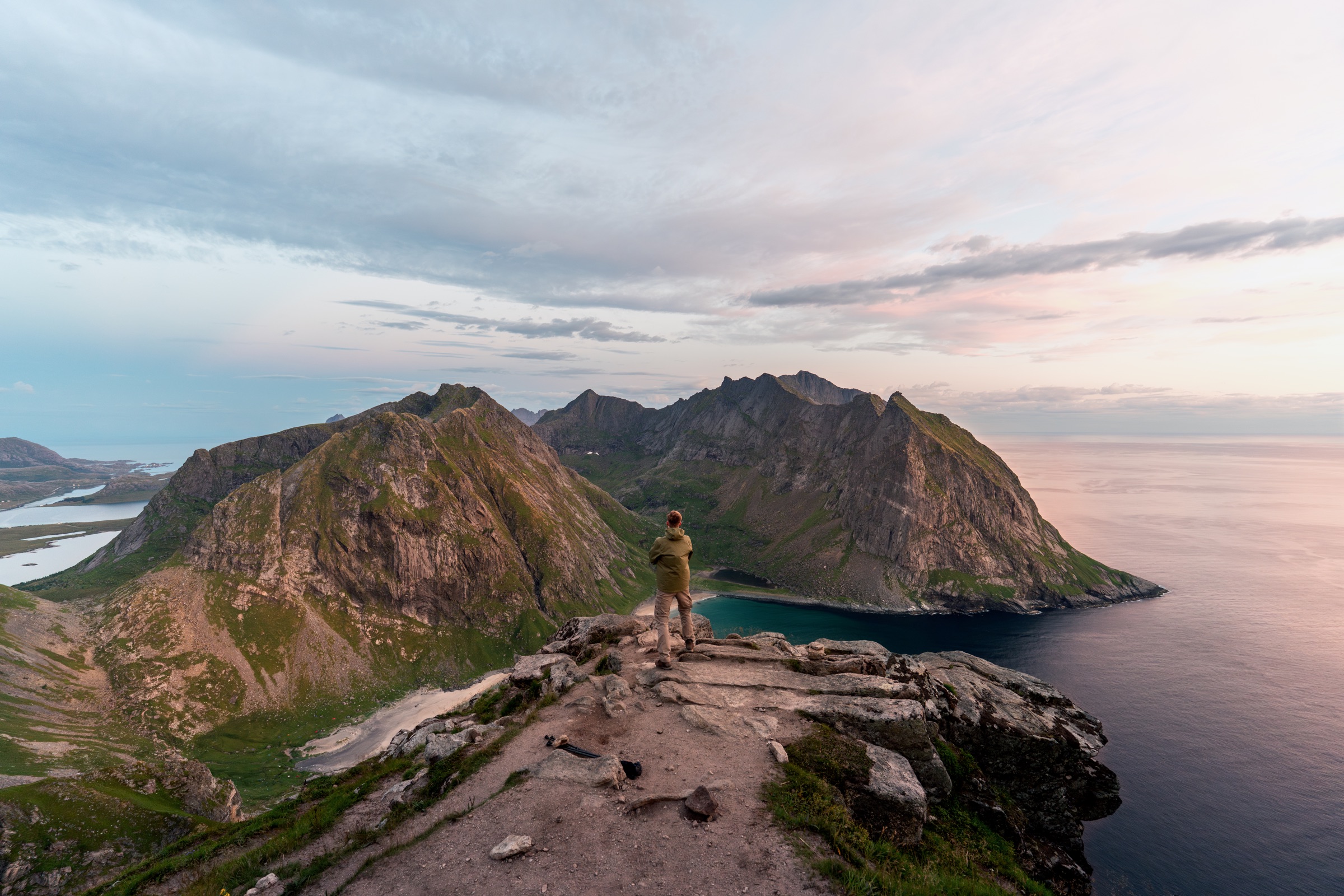 Hiker overlooking Kvalvika Beach, mountain peaks and a fjord at sunset in the Lofoten Islands